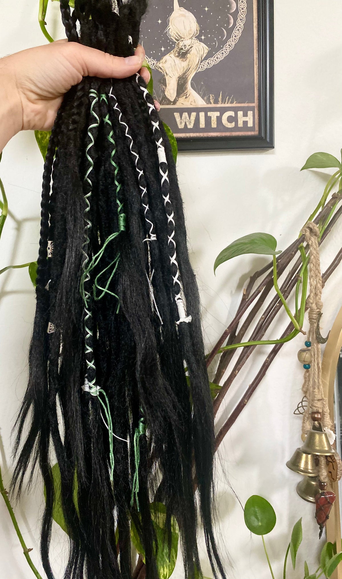 Black synthetic dreadlock extensions with white and green criss-cross decoration. In the background is a photo about a witch with a moon, plants and bells. 