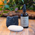 Bottle of Raw Root Hydrating Oil, a bar of shampoo soap bar and detox bomb on a wooden surface with a plant in the background.