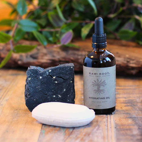 Bottle of Raw Root Hydrating Oil, a bar of shampoo soap bar and detox bomb on a wooden surface with a plant in the background.